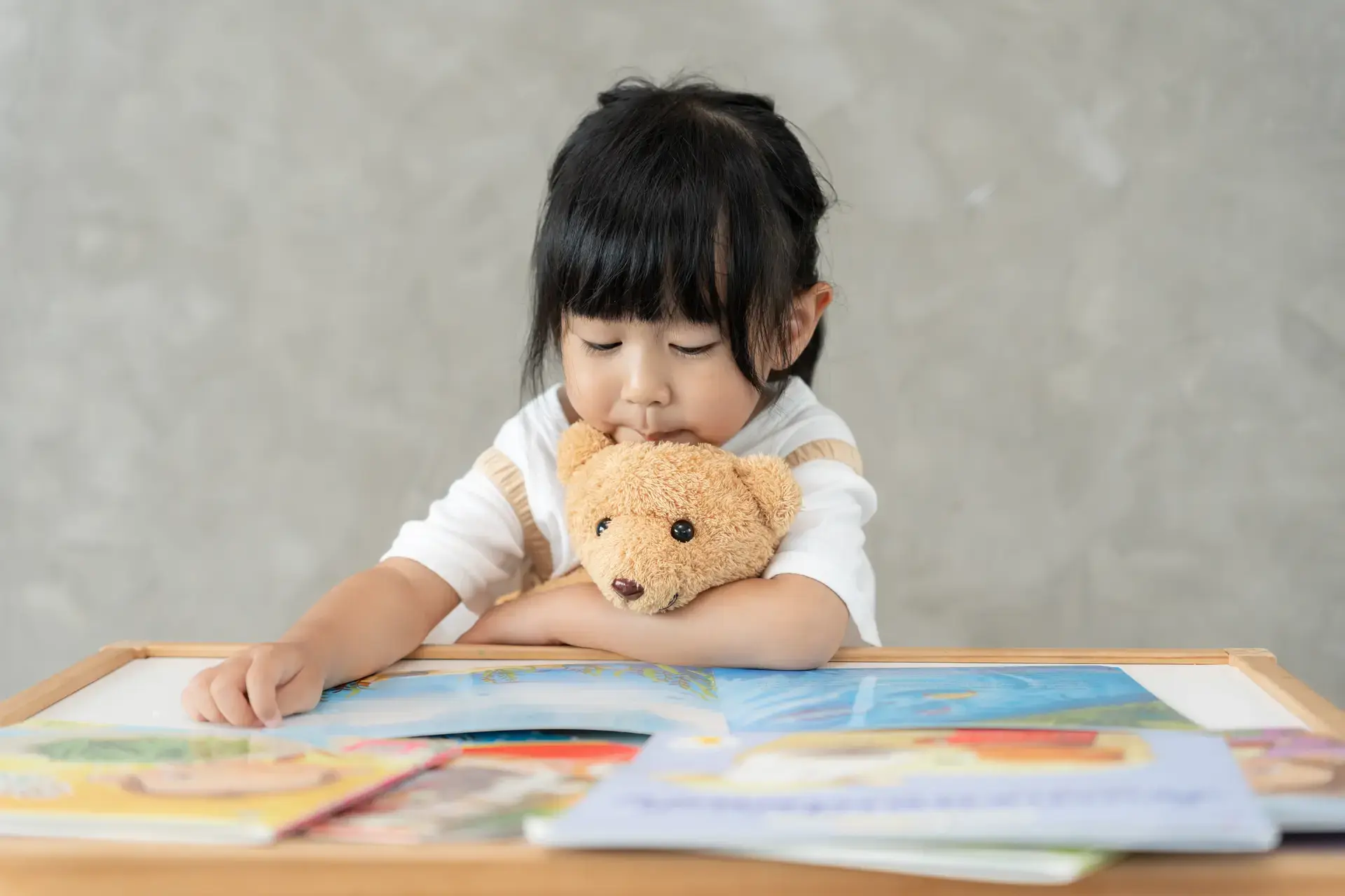 Asyanong batang babae na nagbabasa ng mga libro habang may yakap na teddy bear.